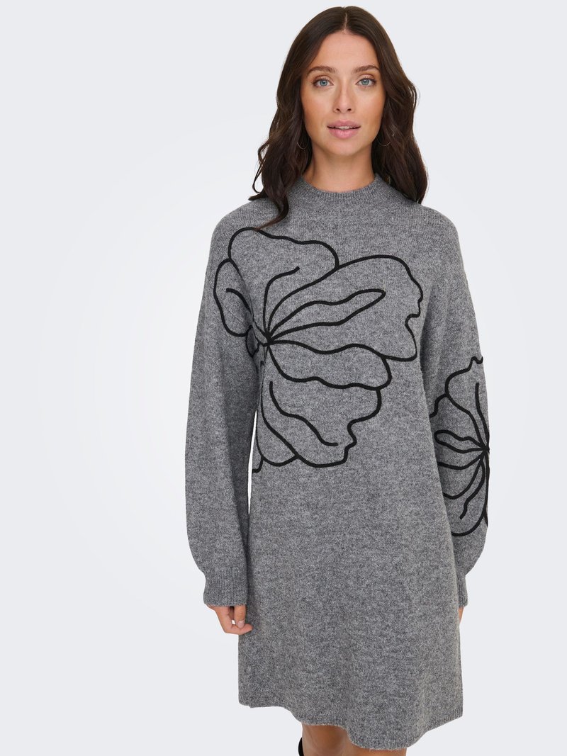 Femme porte une robe en maille grise à manches longues avec de grands motifs floraux abstraits noirs sur le devant et les manches, debout devant un fond uni.