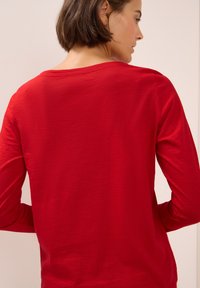 Maglia rossa a maniche lunghe con vestibilità rilassata, scollatura rotonda e tessuto morbido e testurizzato. La vista posteriore mostra una leggera drappeggiatura e dettagli di cucitura sottili.