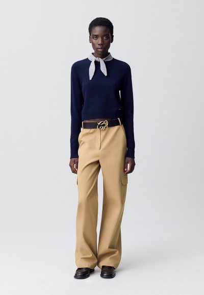 Femme debout portant un pull marine avec une cravate grise, un pantalon beige à large pattes avec des poches latérales, une ceinture noire avec boucle dorée et des chaussures noires.
