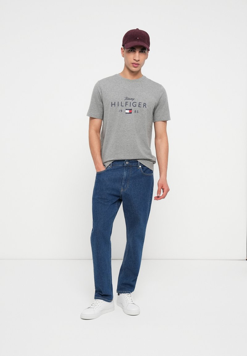 Mand iført en grå Tommy Hilfiger t-shirt, blå jeans, hvide sneakers og en bordeaux cap, stående med den ene hånd i lommen.