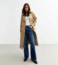 Beige trenchcoat med stora knappar och bälte i midjan, bärs över en vit topp och vida blå jeans. Svarta skor fulländar outfiten.