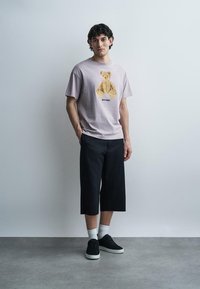 Νεαρός άνδρας στέκεται ανάμεσα στον τοίχο φορώντας ανοιχτό μωβ μπλουζάκι με εκτύπωση αρκουδάκι, μαύρο φαρδύ κοντό παντελόνι, λευκές κάλτσες και μαύρα αθλητικά παπούτσια.