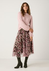 Helles rosa gestricktes Sweatshirt mit langen Ärmeln, kombiniert mit einem transparenten, gemusterten Maxikleid in Rosa- und Grüntönen sowie dunkelgrünen Stiefeletten mit Absatz.