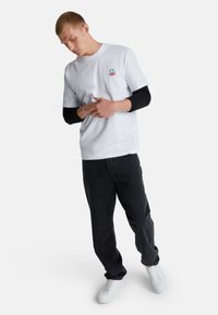 Mladý muž v bílém tričku s malým logem, dlouhý rukáv černého trička pod tím, černých kalhotách a bílých teniskách, stojící se sepnutýma rukama.