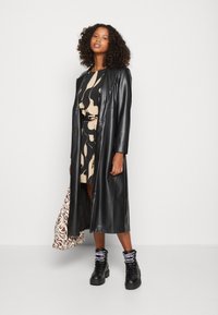 Um casaco longo em couro sintético preto sobre um vestido com padrão abstrato em bege e preto, combinado com botas pretas de cordão e uma mala estampada.