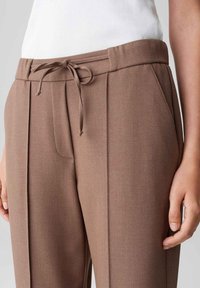 Femme portant un pantalon marron taille haute avec des plis devant, des poches latérales et une ceinture à cordon nouée, associé à un haut côtelé blanc.