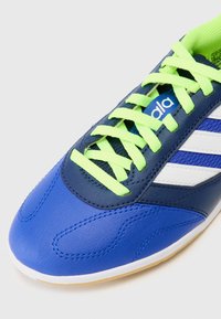 Zapatilla deportiva azul con parte superior de cuero texturizado, rayas blancas y cordones de un verde brillante. Punta redondeada y suela de goma para mayor tracción.