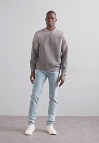 Felpa grigia con collo tondo, polsini e orlo a costine, abbinata a jeans azzurri chiari e sneakers bianche, in piedi davanti a uno sfondo neutro.