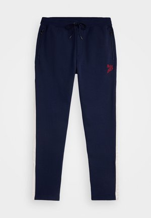 Mørkeblå joggebukser med elastisk midjebånd, snøre, lommer med glidelås og hvite sidelister. Har rødt brodert logo på forsiden.