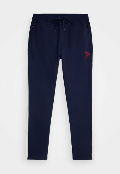 Calças de treino azul-marinho com cintura elástica, cordão para ajuste, bolsos com fecho e riscas brancas laterais. Apresenta um logótipo bordado em vermelho na frente.