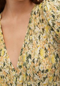 Blumenkleid in Gelb-, Grün- und Weißtönen. Hergestellt aus leichtem Stoff mit V-Ausschnitt und strukturiertem, zerknittertem Muster. Kurze Puffärmel.