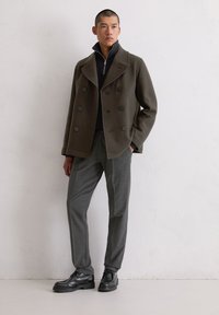 Ciemnozielony dwurzędowy płaszcz typu pea coat z dużymi czarnymi guzikami, w połączeniu z grafitowymi spodniami i czarnymi sznurowanymi butami. Gładka tekstura.