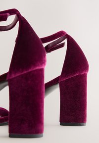 Pár bordó sametových sandálů na podpatku ve tvaru bloku s kotníkovými pásky na jednobarevném světlém pozadí.