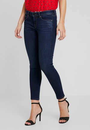 Žena v tmavě modrých úzkých džínách, černých sandálech na vysokém podpatku s páskem kolem kotníku a červeném vzorovaném topu.