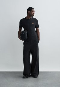 Musta lyhythihainen T-paita, jossa on pieni oranssi brodeeraus, yhdistettynä löysiin mustiin housuihin ja pieneen, pehmeään mustaan laukkuun.