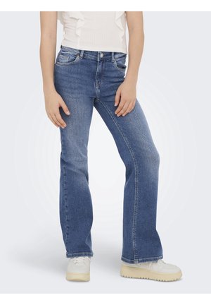 Personne portant un jean bootcut bleu, des baskets blanches et un haut blanc sans manches avec des volants, debout sur un fond clair uni.
