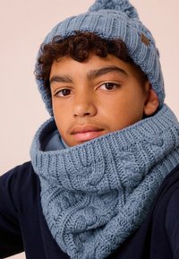 Cappello lavorato a maglia di colore azzurro chiaro con pom-pom e sciarpa a collo coordinata, caratterizzata da un motivo a treccia texture. Entrambi sono spessi e accoglienti.