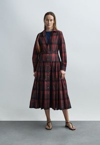 Raudonos ir tamsiai languotos suknelės su ilgomis rankovėmis, sagomis priekyje ir sluoksniuota sijonu. Derinama su juodais šlepetėmis ir ruda rankine.