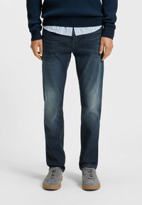 Dark blue jeans with a straight leg design, slight fading on the thighs, worn with a light blue striped shirt and grey sneakers.