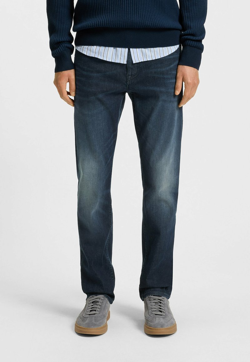 Dark blue jeans with a straight leg design, slight fading on the thighs, worn with a light blue striped shirt and grey sneakers.