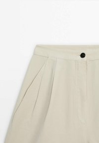 Pantalon beige taille haute avec plis devant, poches latérales, fermeture à bouton noir et ceinture en tissu.