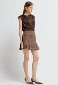 Top en satin marron avec des épaules structurées, associé à une mini-jupe plissée en tweed dans des tons bruns, ornée de détails dorés et de talons fins.