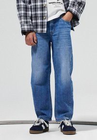 Jean en denim bleu clair avec une coupe droite, présentant des détails de décoloration subtils. Associé à une chemise à carreaux noire et blanche et des baskets.