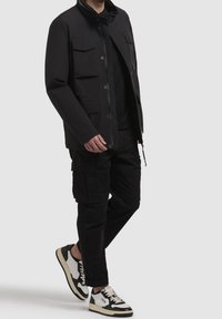Homme portant un pantalon cargo noir, une veste noire avec des poches à rabat et des baskets noir et blanc, debout devant un fond gris uni.
