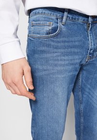 Jeans de mezclilla azul claro con corte ajustado, que cuentan con cinco bolsillos, un sutil desgaste y detalles de costura a lo largo de las costuras.