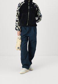 Černá fleecová bunda s vzorem a vysokým límcem, spárovaná s tmavě modrými kalhotami. Modelka drží béžovou tašku. Bílé tenisky dokončují outfit.