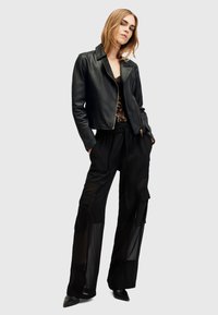 Giacca cropped in pelle nera con zip frontale, indossata sopra un top in pizzo. Pantaloni neri trasparenti a gamba larga con tasche applicate e tessuto testurizzato.