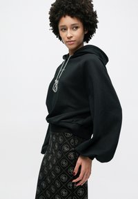 Schwarzer Kapuzenpullover mit Kordelzug, ausgestattet mit weiten Ärmeln. Kombiniert mit einem gemusterten schwarzen Rock mit geometrischen Mustern.