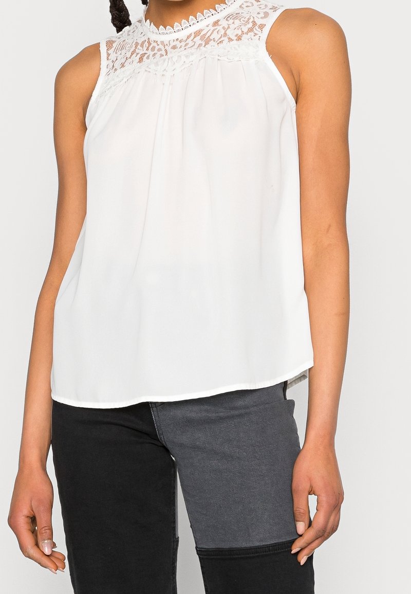 Vit ärmlös topp med spettsyning och ett luftigt tyg. Rundad nederkant, lätt textur och diskret drapering vid halsringningen.