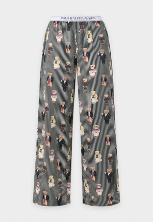 Femme portant un crop top bleu marine avec le texte "POLO" et un pantalon de pyjama rayé avec des illustrations d'ours, debout pieds nus, mains dans les poches.