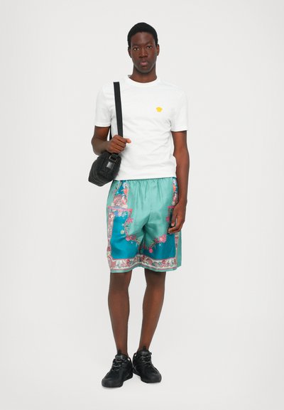 Vit kortärmad skjorta med ett gult emblem, teal färgade shorts med blommönster, svarta sneakers och en svart väska.