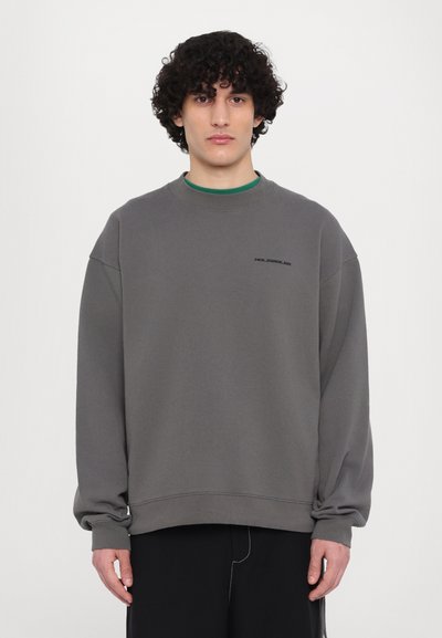 Grå oversized hettegenser i myk stoff med rund hals, senket skuldre, og en liten svart logodetalj på brystet.