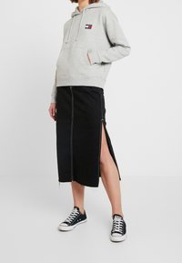 Sudadera gris con capucha y bolsillo frontal, con un logo, combinada con una falda de mezclilla negra con aberturas laterales y costuras blancas. Se lleva con zapatillas negras.