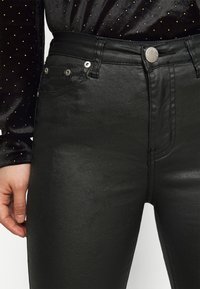 Jeans pretos de cintura alta feitos de um material liso e brilhante. As características incluem um fecho frontal com botão e dois bolsos traseiros com costuras visíveis.