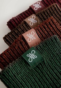 Adītas galvassegas tumši sarkanā, brūnā, oranžsarkanā un zaļā krāsā, ar strukturētu rakstu un mazu apzīmējuma auduma birku ar balto izšuvumu.