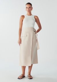 Bleda bež krilna krila z stransko žepom in pasom, v kombinaciji z prilegajočim belim rebrastim topom. Model nosi črne minimalistične sandale.