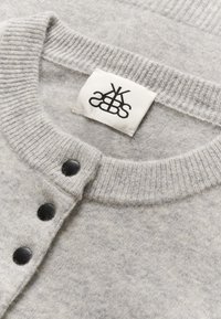 Harmaa neulottu neuletakki, jossa on resorinen pääntie, mustat napit edessä ja sisäpuolella ommeltu kangasmerkki logolla.