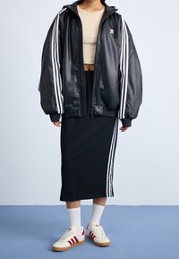 Person, die eine schwarze Adidas-Jacke und einen Rock mit weißen Streifen, ein beiges Crop-Top, weiße Socken und weiße Sneaker mit roten Streifen trägt.