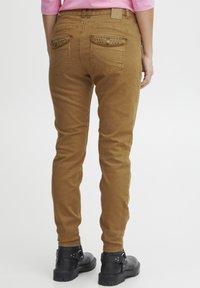 Brown denim pants with a fitted design, featuring studded detailing on the back pockets and a smooth texture. Black ankle boots worn.