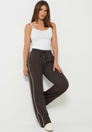 Vrouw staat tegen een witte muur, draagt een wit hemdje, zwarte broek met trekkoord en witte zijstrepen, en witte sneakers.
