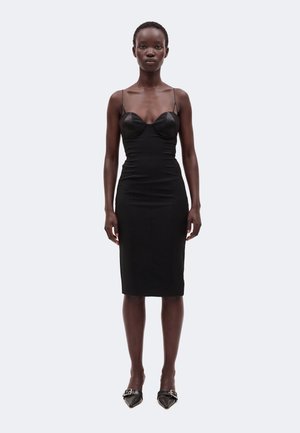 Femme debout face à l’avant, portant une robe midi noire ajustée à fines bretelles et des ballerines noires pointues avec des boucles.