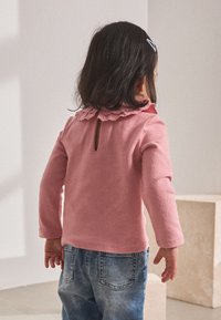 Langarmige roze trui met een golvende halslijn en een keyhole-detail aan de achterkant, gecombineerd met blauwe spijkerbroeken. Zachte, gestructureerde stof.