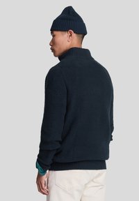 Suéter de punto azul oscuro con un cuello alto, textura acanalada, mangas largas y un corte relajado. Llevado con un gorro a juego.