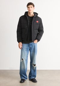 Giubbotto piumino nero con etichetta rossa, dotato di cappuccio e polsini elasticizzati, indossato sopra una maglietta nera e abbinato a jeans in denim blu strappati.