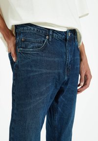 Vaqueros de mezclilla azul oscuro con diseño de pierna recta, bolsillos delanteros y herrajes metálicos. La tela presenta una ligera textura y detalles de desgaste.