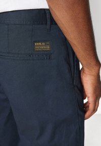 Tamno plave hlače glatke teksture, s vidljivim džepom i brendiranim logom s zlatnim tekstom.